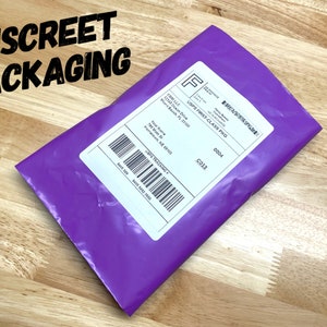 Op de afbeelding: Een paarse gevoerde envelop met een wit verzendlabel. Het label heeft de tekst "USPS First Class PKG" en een track-and-trace nummer. De envelop ligt op een houten oppervlak.
