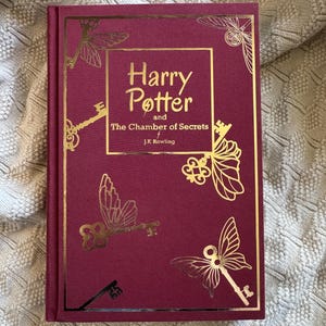 May include: A hardcover book titled "Harry Potter and The Chamber of Secrets" by J.K. Rowling. The book has a burgundy cover with gold foil accents, including keys and butterfly-key designs. The title is in gold lettering.