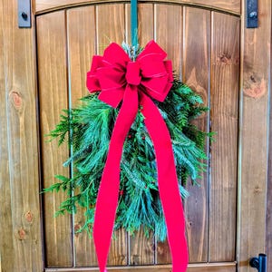 May include: A teardrop-shaped evergreen swag featuring a mix of pine and cedar needles with small red berries. A large, vibrant red velvet bow is secured at the top, with two long, flowing ribbon tails extending downward over the greenery.