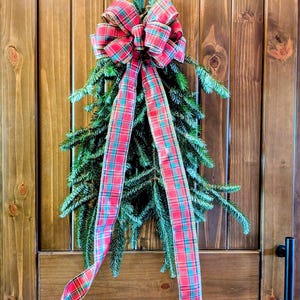 May include: A teardrop-shaped evergreen swag made of dark green pine branches, topped with a large, multi-looped bow featuring a red, green, and blue plaid pattern. Two long, matching plaid ribbon tails hang down from the center of the bow.