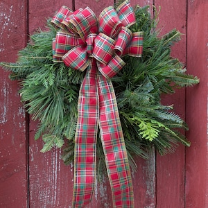 May include: A circular wreath constructed from dense green evergreen boughs, including pine and cedar needles. A large, multi-looped bow made of red, green, and gold plaid ribbon is attached to the top, with two long ribbon tails hanging down.