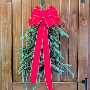 May include: A festive evergreen swag featuring a large, vibrant red velvet bow with silver-trimmed edges. The lush, deep green pine branches are arranged in a downward-pointing teardrop shape, suspended by a green metal hook on a wooden door.