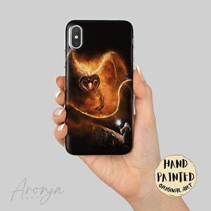 May include: A black smartphone case featuring a detailed illustration of a fiery, winged creature with glowing orange and yellow flames. Below the creature, a small figure stands on a dark ledge holding a glowing white staff. The art is vibrant.