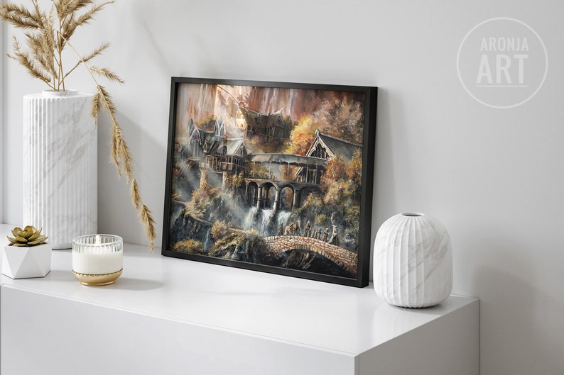 Op de afbeelding: Een ingelijste rechthoekige kunstdruk met een gedetailleerd landschapsschilderij van een nederzetting in elfenstijl met stenen bruggen, watervallen en herfstkleurig gebladerte. Het kunstwerk zit in een dunne zwarte lijst en toont de tekst ARONJA ART.