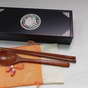 Puede incluir: Una caja de madera negra con detalles plateados en las esquinas y una incrustación de nácar. Una cuchara de madera, palillos y una pequeña bolsa naranja con elementos decorativos se muestran frente a la caja. La cuchara y los palillos son marrones.