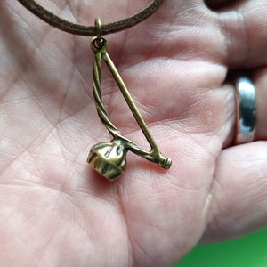 Puede incluir: Un collar con un colgante de color dorado que representa un berimbau, un instrumento musical tradicional brasile&ntilde;o. El colgante est&aacute; suspendido de un cord&oacute;n de cuero marr&oacute;n.