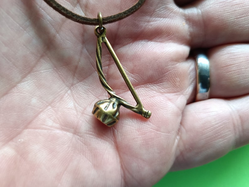 Puede incluir: Un collar con un colgante de color dorado que representa un berimbau, un instrumento musical tradicional brasile&ntilde;o. El colgante est&aacute; suspendido de un cord&oacute;n de cuero marr&oacute;n.