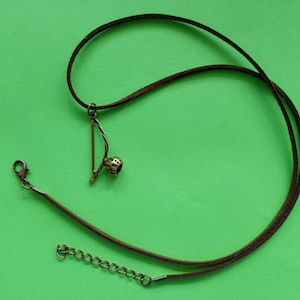 Puede incluir: Un collar de cuero marr&oacute;n con un peque&ntilde;o colgante dorado en forma de arco y flecha. El colgante tiene una piedra redonda oscura en el centro.