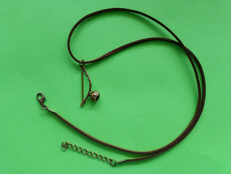 Puede incluir: Un collar de cuero marr&oacute;n con un peque&ntilde;o colgante dorado en forma de arco y flecha. El colgante tiene una piedra redonda oscura en el centro.