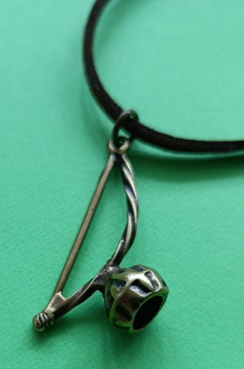 Puede incluir: Un collar de cord&oacute;n de cuero marr&oacute;n con un colgante de bronce con forma de berimbau, un instrumento musical brasile&ntilde;o.