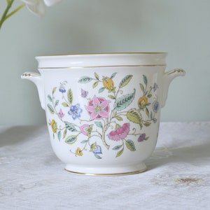 Puede incluir: Un macetero de cerámica blanca con un diseño floral y ribete dorado. El macetero tiene dos asas y está sentado sobre un mantel blanco. Una orquídea blanca está en un jarrón detrás del macetero.