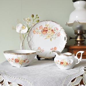 Pode incluir: Um conjunto de chá de porcelana branca com desenhos florais, incluindo uma tigela, um prato e um cremeira. O conjunto tem acabamentos dourados e é exibido sobre uma toalha de mesa de renda branca. Uma lâmpada a óleo e um vaso com flores brancas também estão presentes.