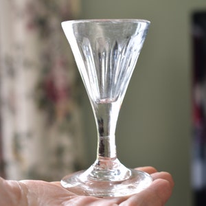 Puede incluir: Un vaso de chupito de vidrio transparente con un diseño acanalado y un tallo largo. El vaso está sentado en una mano.