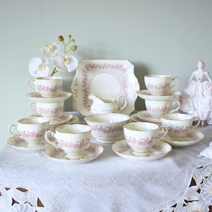 Könnte beinhalten: Ein Vintage-Teeservice mit rosa und blauen Blumenmustern. Das Set umfasst Tassen, Untertassen, einen Servierteller, einen Sahnekännchen und eine Zuckerdose. Das Teeservice ist auf einer weißen Tischdecke mit floraler Spitzenborte angeordnet.