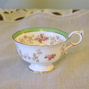 May include: A white teacup with a green rim and floral pattern. The cup has a delicate handle and a pedestal base.
