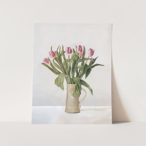 May include: A bouquet of pink tulips in a cream-colored ceramic pitcher with gold accents. The tulips are arranged in a vase on a white surface.