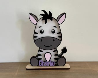 May include: A wooden zebra figurine with black and white stripes, a black mane, and the name "Charlie" in purple. The zebra has pink ears and a pink heart-shaped design on its chest. It sits on a wooden base.