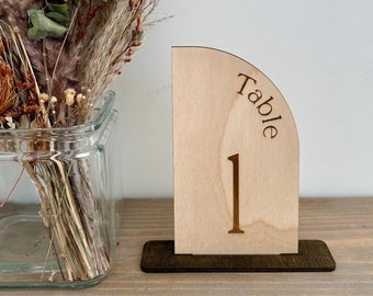 May include: A wooden table number with a rounded top, featuring the text "Table 1" in a brown font. The number is displayed on a wooden shelf with a vase of dried flowers in the background.