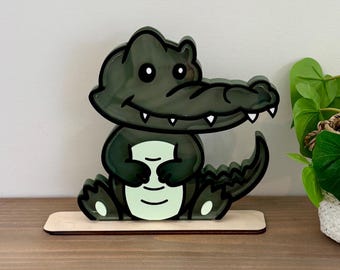 May include: A green and black cartoon alligator figurine with a toothy grin. The figurine is made of wood and has a light green glow. It is sitting on a wooden base.