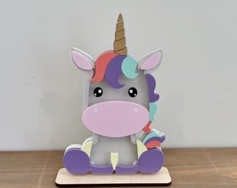 May include: A whimsical unicorn figurine with a light gray body, a pink muzzle, and a multicolored mane and tail. The unicorn has a golden horn and is seated on a wooden base. The figurine is a decorative item.