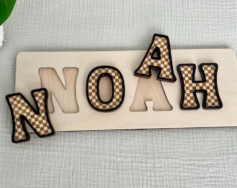 May include: Wooden puzzle with the name NOAH spelled out in brown and white checkered letters. The letters are separate and can be placed in the puzzle slots.