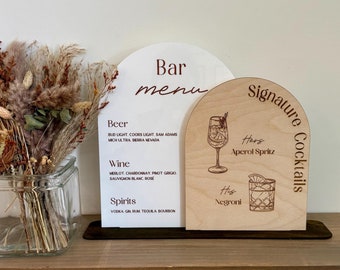 May include: A bar menu and signature cocktail signs. The bar menu is white with brown text, listing beer, wine, and spirits. The cocktail sign is wood with Aperol Spritz and Negroni illustrations.