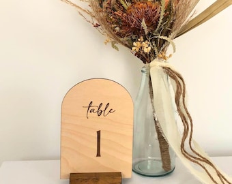 May include: Wooden table number with the text "table 1" engraved on it. The number is displayed in front of a bouquet of dried flowers in a clear glass vase.