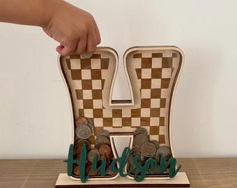 May include: Wooden letter "H" shaped coin bank with a checkerboard pattern. The bank is filled with coins and has the name "Hudson" in green script at the base. A hand is placing a coin into the bank.