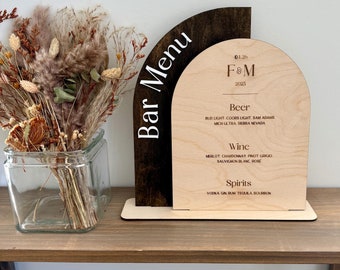 May include: A wooden bar menu sign with a brown and light wood arch design. The sign lists the drinks available at the bar, including beer, wine, and spirits. The text on the sign reads "Bar Menu", "F&M", "04.26.2025", "Beer", "Bud Light, Coors Light, Sam Adams, Mich Ultra, Sierra Nevada", "Wine", "Merlot, Chardonnay, Pinot Grigio, Sauvignon Blanc, Rose", "Spirits", "Vodka, Gin, Rum, Tequila, Bourbon".