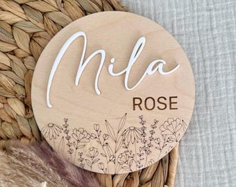 May include: A round wooden sign with the name "Mila Rose" written in white raised letters. The sign is decorated with a floral design around the edge.