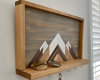 May include: Wooden key holder with a mountain design and a shelf for sunglasses. The mountain design is made of different colored wood pieces. There are five hooks on the bottom of the shelf.