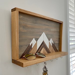 May include: Wooden key holder with a mountain design and a shelf for sunglasses. The mountain design is made of different colored wood pieces. There are five hooks on the bottom of the shelf.
