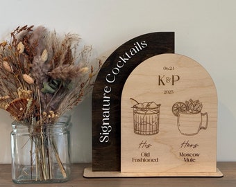 May include: A wooden sign with a brown and light wood finish. The sign is shaped like an arch and has the text "Signature Cocktails" on the left side. The right side of the sign has the text "06.24 K&P 2025" and two illustrations of cocktails. The text "His Old Fashioned" is below the first illustration and the text "Hers Moscow Mule" is below the second illustration.