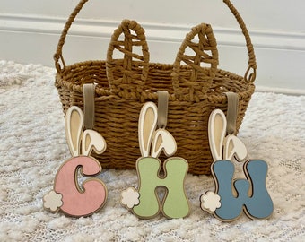 May include: Three wooden bunny-shaped ornaments with the letters C, H, and W painted in pink, green, and blue. The ornaments are hanging from a wicker basket with bunny ears.