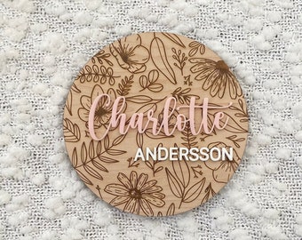 May include: A round wooden sign with a floral design and the name "Charlotte Andersson" written in pink and white lettering.