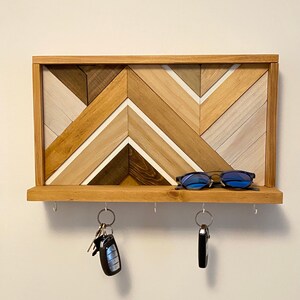 May include: A wooden key holder with a geometric chevron pattern in shades of brown, white, and gray. The key holder has a shelf with four hooks for hanging keys and other items. Sunglasses are resting on the shelf.