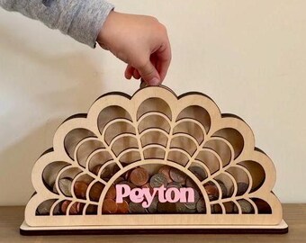 May include: Wooden coin bank shaped like a half-circle with a scalloped edge. The bank has a cutout design and is filled with coins. The name "Peyton" is written in pink letters on the front.