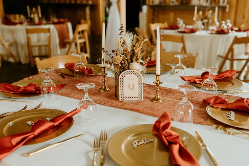 Peut inclure: Une table dress&eacute;e avec des assiettes dor&eacute;es, des serviettes rouges et des verres transparents. Un num&eacute;ro de table en bois avec le num&eacute;ro "9" est au centre de la table. La table est d&eacute;cor&eacute;e d'un chemin de table et d'une composition florale.