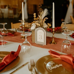 Peut inclure: Une table de mariage avec des assiettes dor&eacute;es, des serviettes rouges et de la verrerie transparente. Un porte-num&eacute;ro de table en bois avec le num&eacute;ro "3" est au centre de la table. La table est d&eacute;cor&eacute;e d'un arrangement floral dans une bo&icirc;te en bois.