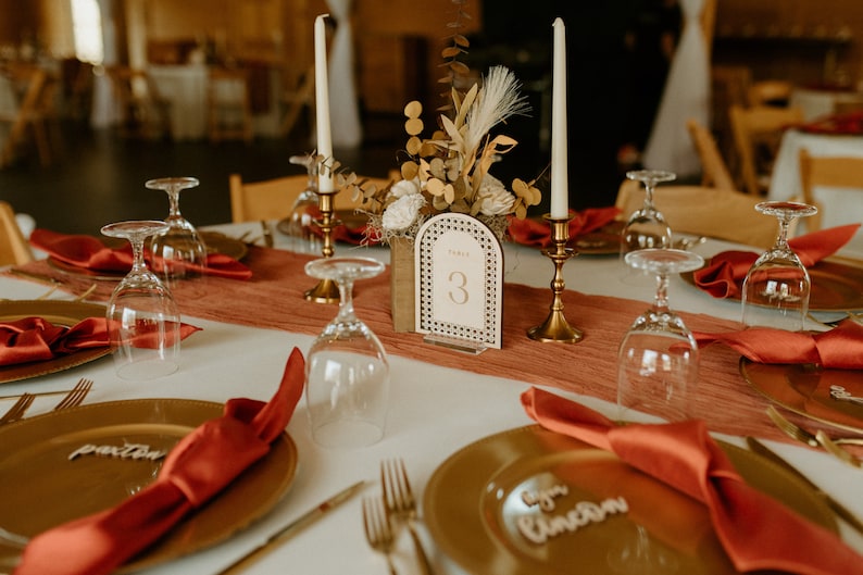 Peut inclure: Une table de mariage avec des assiettes dor&eacute;es, des serviettes rouges et de la verrerie transparente. Un porte-num&eacute;ro de table en bois avec le num&eacute;ro "3" est au centre de la table. La table est d&eacute;cor&eacute;e d'un arrangement floral dans une bo&icirc;te en bois.
