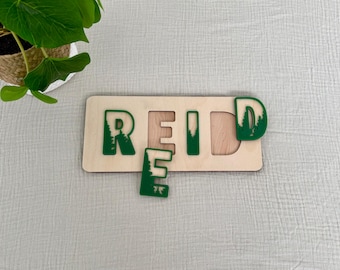 May include: Wooden puzzle with the name "REID" spelled out in green letters with a forest design. The letters are separate pieces that fit into the puzzle board.