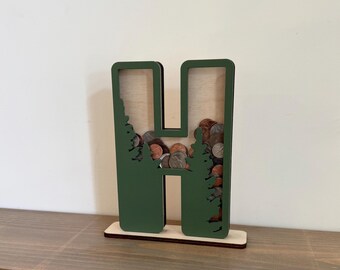 May include: A green wooden letter H shaped coin bank with a cutout design of trees. The bank is filled with coins.