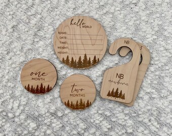 May include: Wooden baby milestone discs with a forest theme. The discs feature the words "one month", "two months", and "hello world". There is also a wooden hanger with the words "NB newborn" and "NB 3".