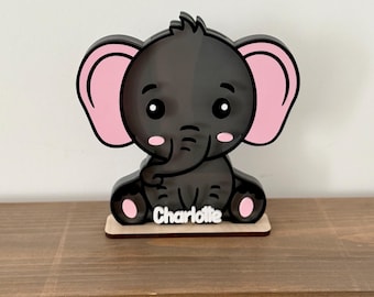 May include: A black and pink elephant-shaped decoration with the name "Charlotte" on a wooden base. The elephant has large pink ears, a trunk, and rosy cheeks. It is sitting on a wooden shelf.