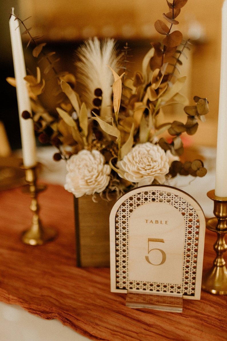Peut inclure: Un num&eacute;ro de table en bois avec un motif tiss&eacute;, comportant le texte "TABLE 5" en lettres dor&eacute;es. Le num&eacute;ro est pr&eacute;sent&eacute; sur un support en acrylique transparent et est entour&eacute; d'un arrangement floral avec des fleurs s&eacute;ch&eacute;es et de la verdure.