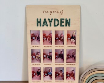 May include: A wooden photo display with a green inscription "one year of HAYDEN" and twelve framed photos of a baby boy at different ages from one month to one year old.
