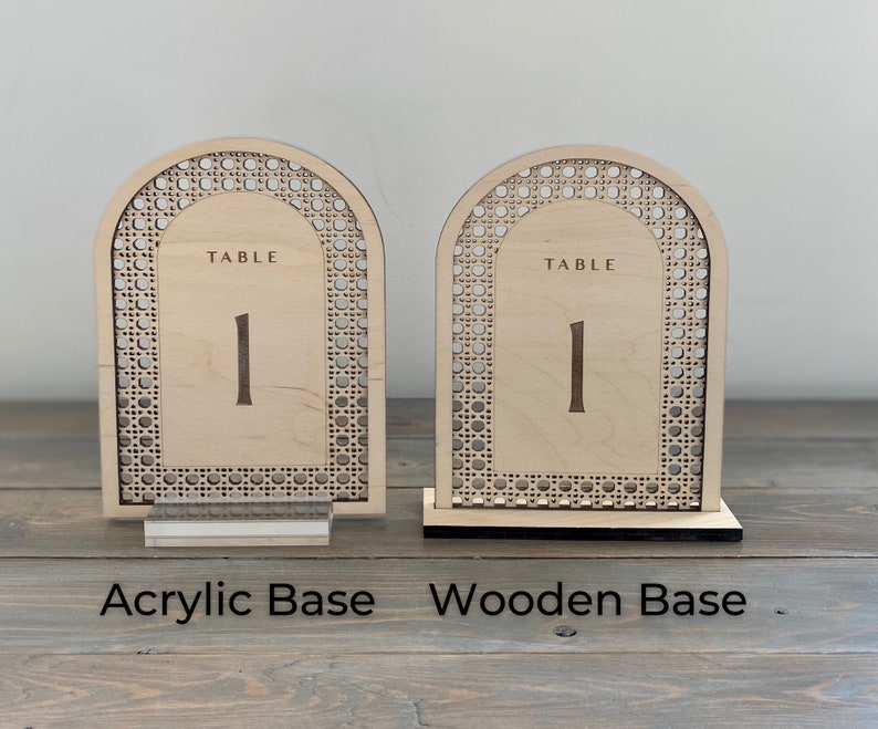 Peut inclure: Deux porte-num&eacute;ros de table en bois avec un motif tiss&eacute;. Le porte-num&eacute;ro de gauche a une base en acrylique et le porte-num&eacute;ro de droite a une base en bois. Les deux porte-num&eacute;ros portent le texte "TABLE 1".