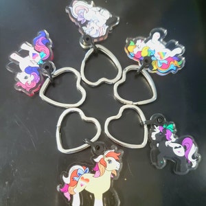 Puede incluir: Seis llaveros de plástico coloridos con ponis de dibujos animados con crines y colas arcoíris. Cada llavero está unido a un anillo de corazón plateado.