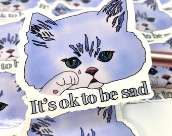 May include: A sticker featuring a cartoon blue cat with a tear rolling down its cheek. The cat is holding its paw to its face and the text "It's ok to be sad" is written below the cat.