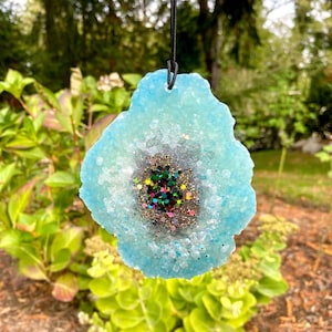 May include: A blue resin pendant with a glittery, iridescent center, resembling a geode. The pendant is hanging from a black cord.